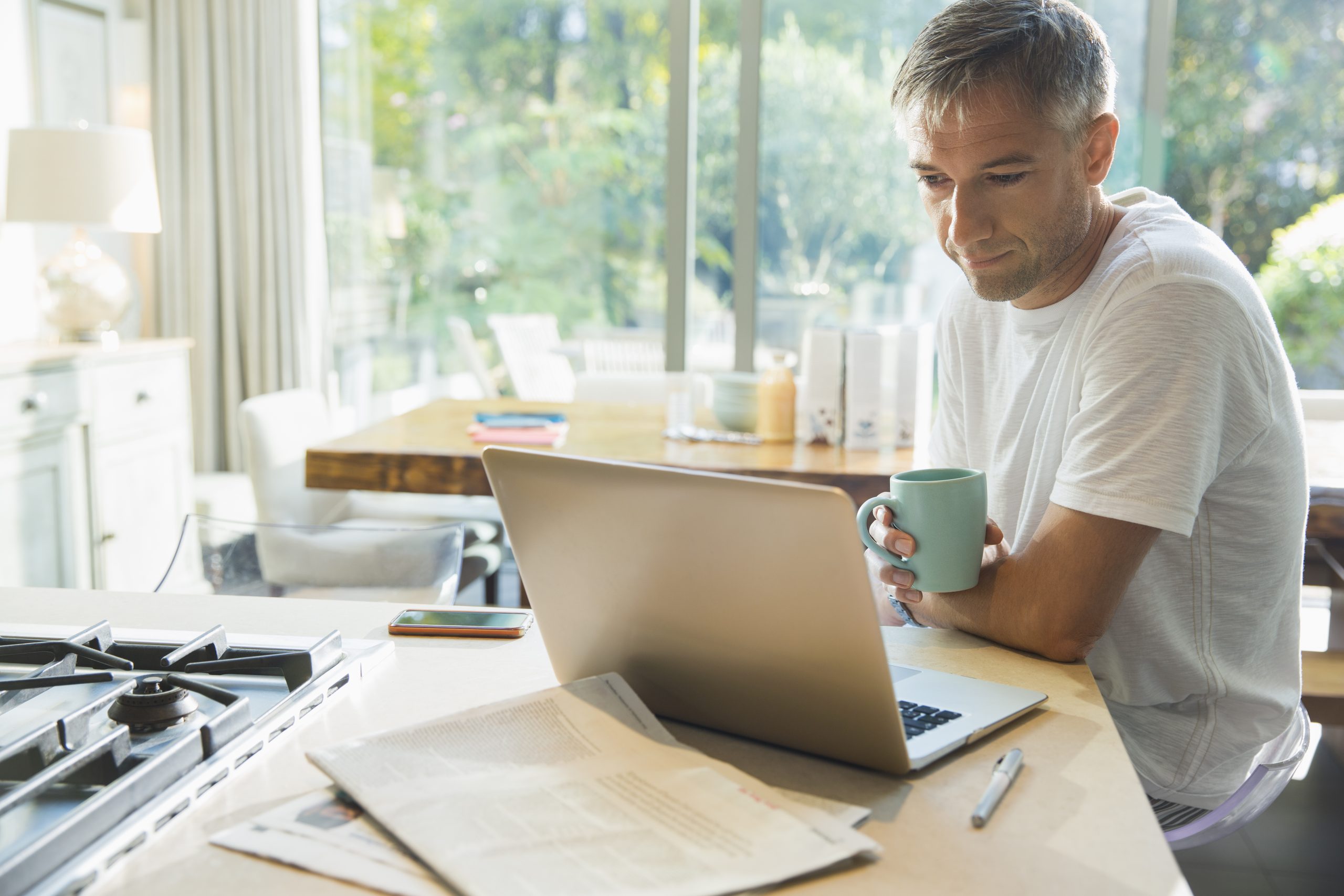 Mann sitzt in der Küche mit einer Tasse Kaffee in der Hand vor seinem Laptop